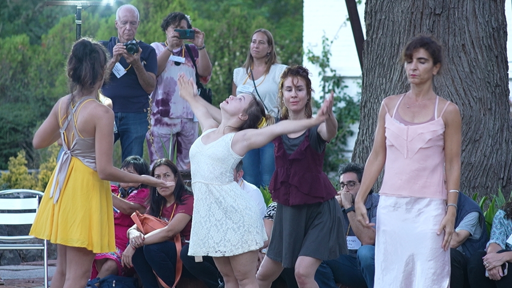 grupo de 7 mujeres bailando vestidas con diferentes colores, una de ellas tiene sindrome de Down.
Hay gente alrededor mirando y sacando fotos